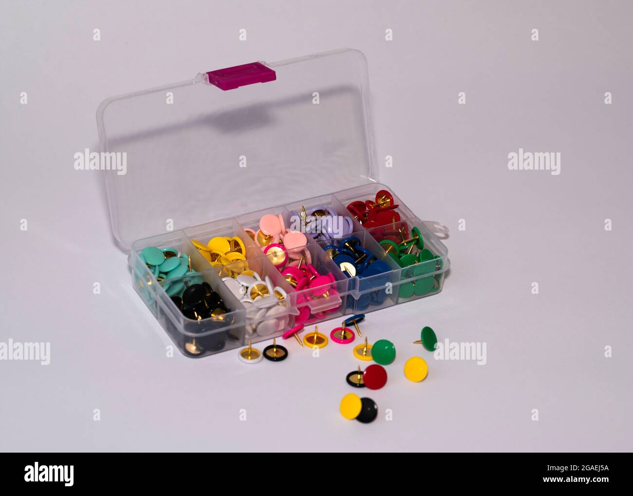 open plastic container with ten different coloured drawing pins in separate dividers with a few scattered pins on a white background Stock Photo