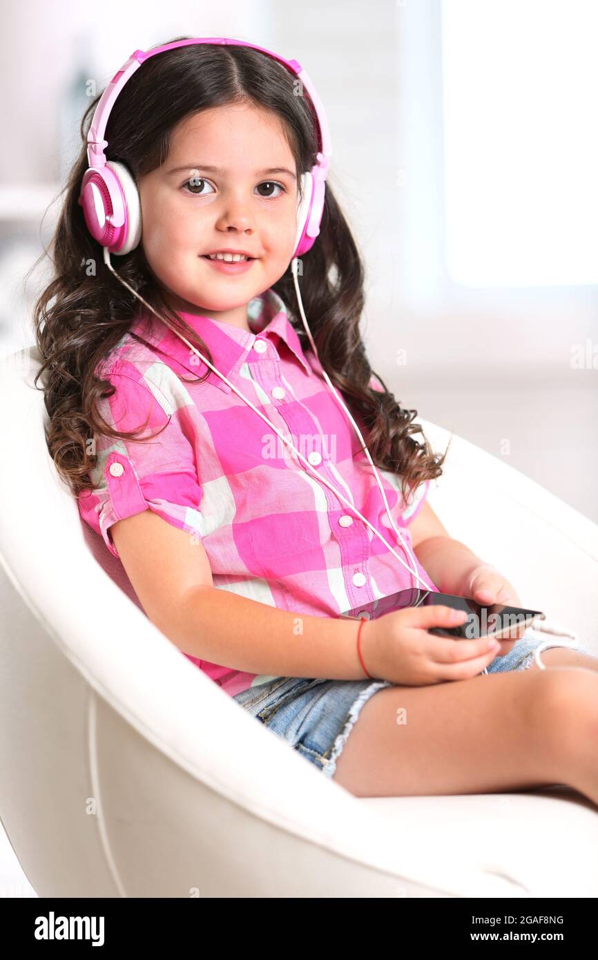 Attractive little girl sitting on comfortable chair and listening music with pink headphones in