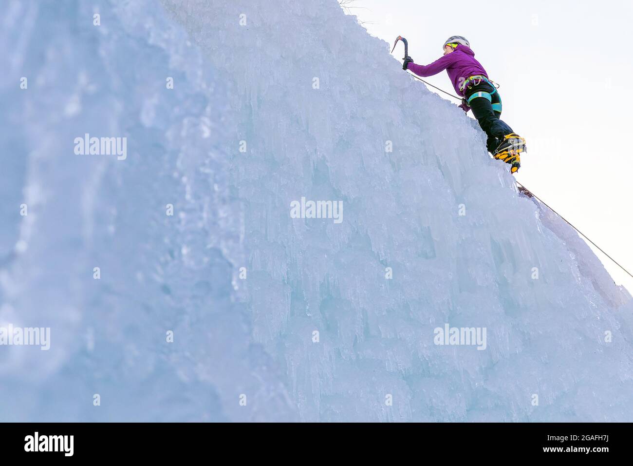 Female ice climber hiking a frozen waterfall, pushing axe pick into the slope and moving up to the top Stock Photo