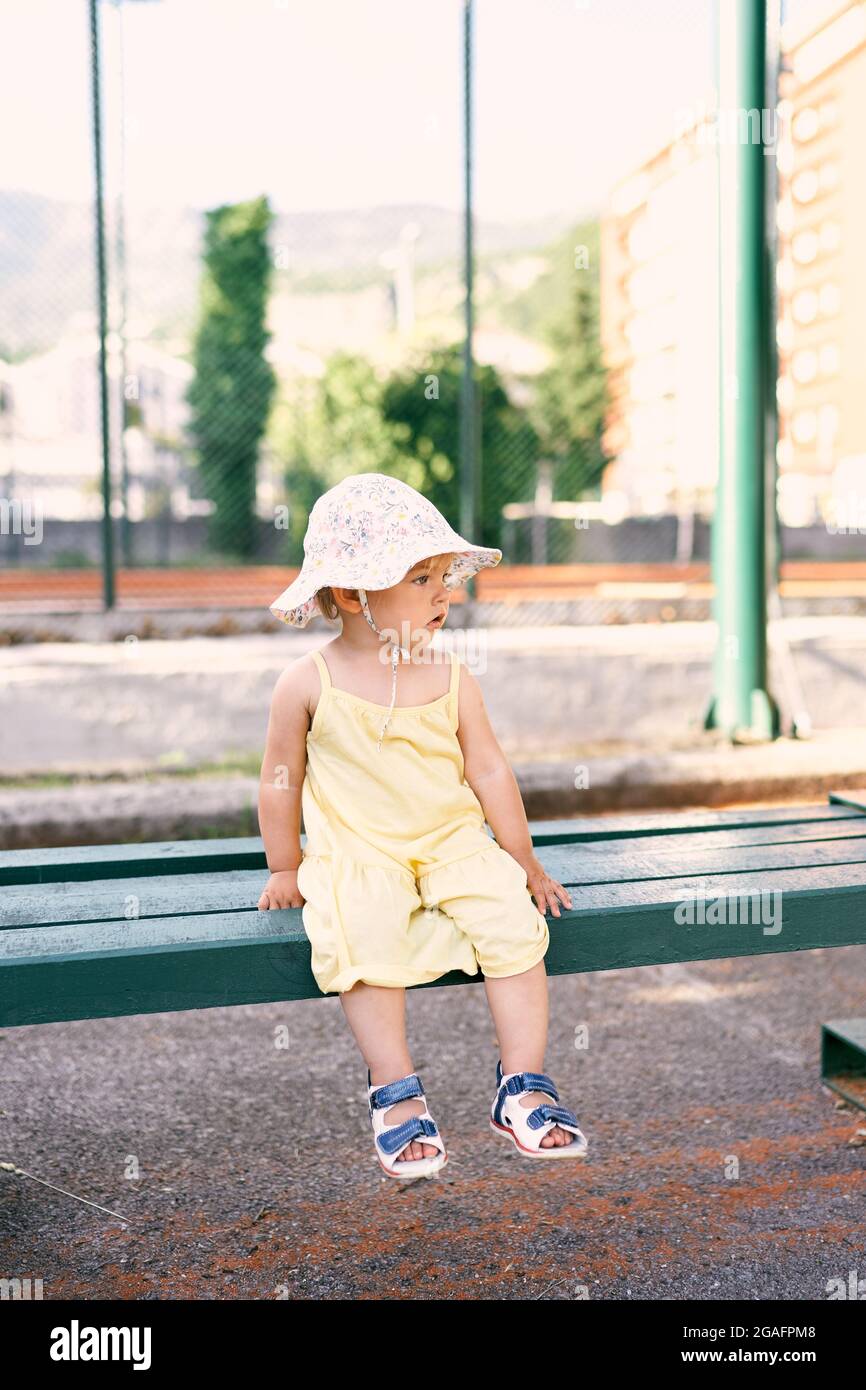 Little girl in a hat sits on a wooden bench near the fence of the park ...