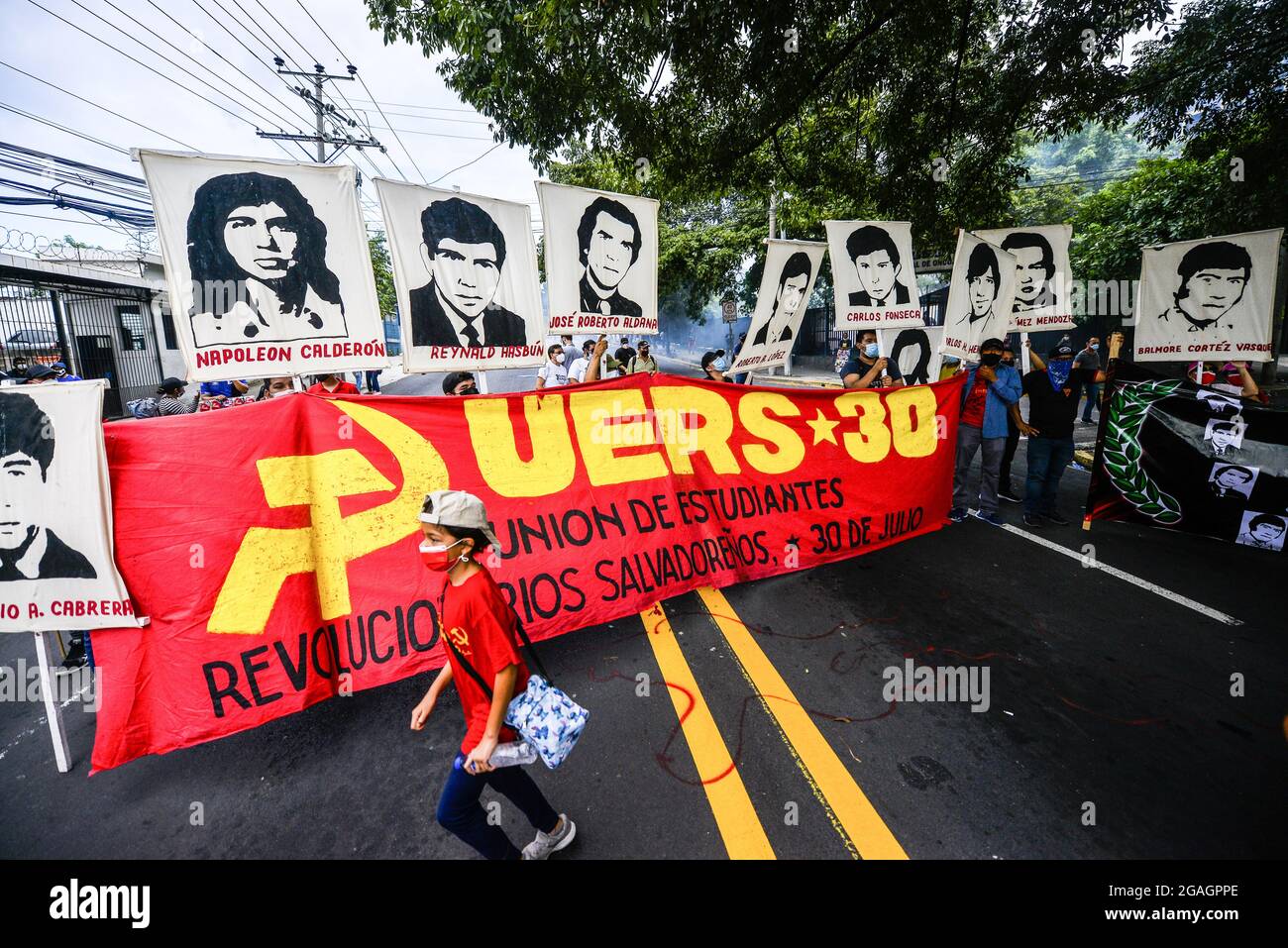 San Salvador, El Salvador. 30th July, 2021. A kid runs in front of a display of a banner and portraits of students killed by the military during a protest in 1975. Students from El Salvador's National University protest against dictatorship during a commemorative march on the 46th anniversary of a government led massacre against students. (Photo by Camilo Freedman/SOPA Images/Sipa USA) Credit: Sipa USA/Alamy Live News Stock Photo