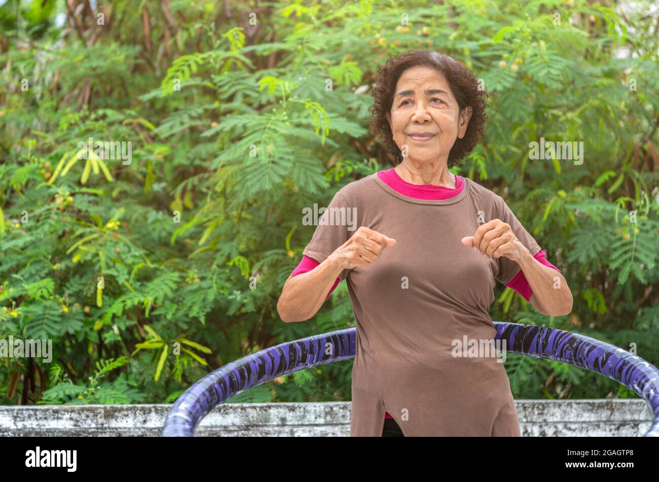 Portrait of healthy Asian senior woman is working out by hula hooping on the terrace of house, background top part of big tree, eyes looking to the bl Stock Photo