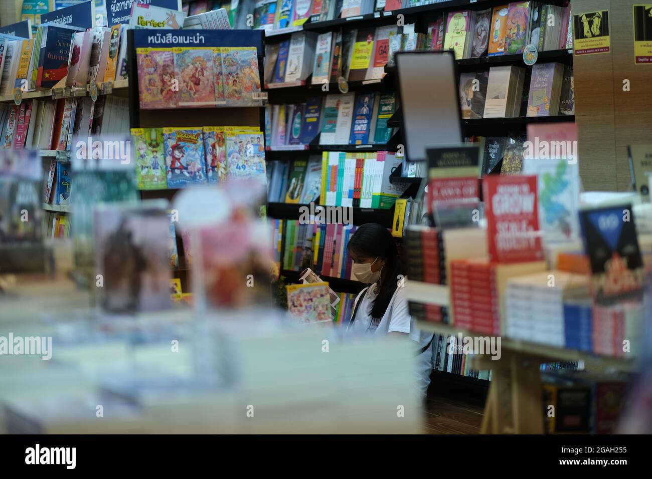 Young Thai woman is reading a book in a comfort of heavy bookshelfs of public library or a bookstore Stock Photo