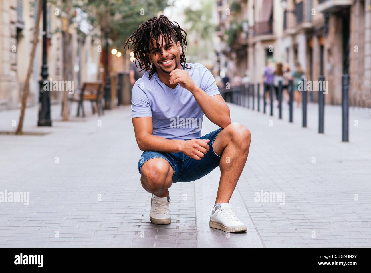 horizontal portrait of a trendy young latin man crouched down. He is in the streets of barcelona and smiles to camera Stock Photo