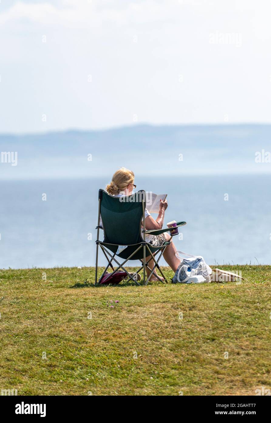 middle-aged lady at the seaside reading a book, older lady sitting at the beach in a chair with a good book, lady enjoying the sunshine and reading. Stock Photo