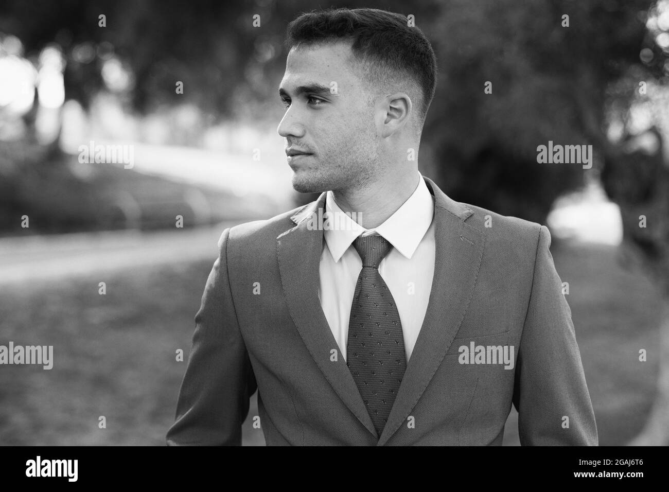 Confident young man in full suit, adjusting sleeve and looking outside, with garden in background Stock Photo