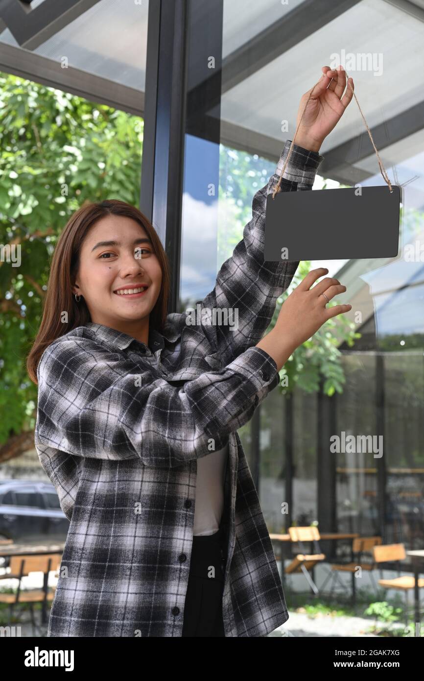 Smiling woman hanging blank wooden sign board on glass door. For your advertising text. Stock Photo