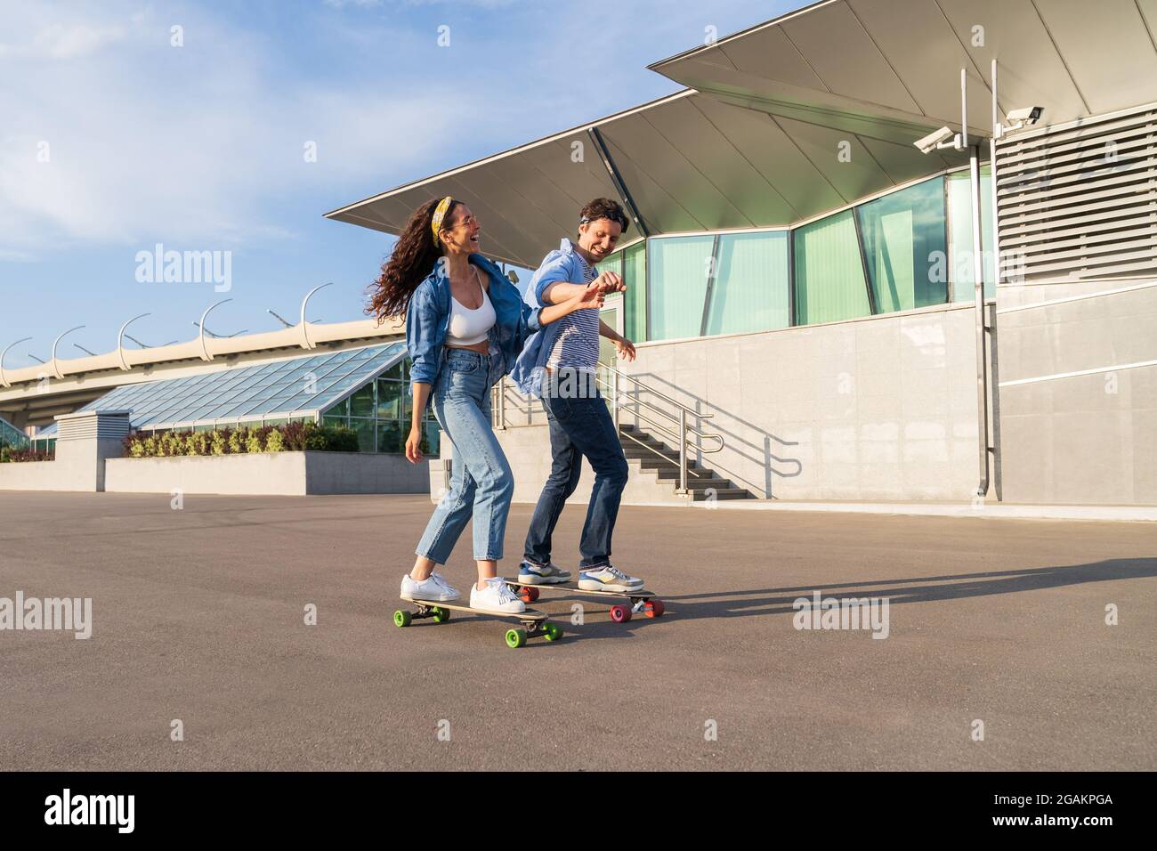Joyful couple on date longboarding together: hipster guy skater teach laughing woman ride skateboard Stock Photo