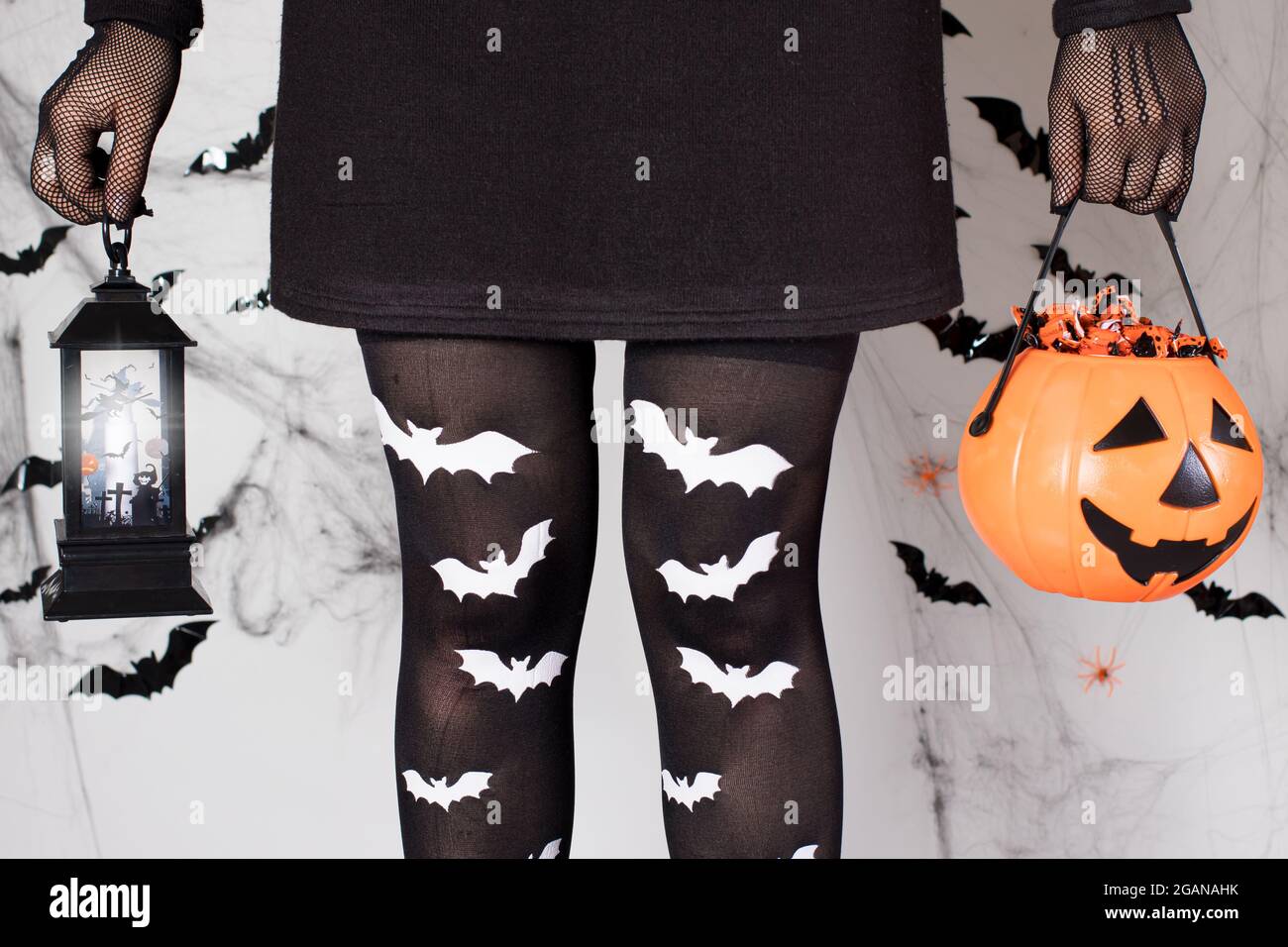 A Halloween girl in a witch costume in bat socks holds a glowing lantern and a pumpkin bucket of candy on a white background with cobwebs. Stock Photo