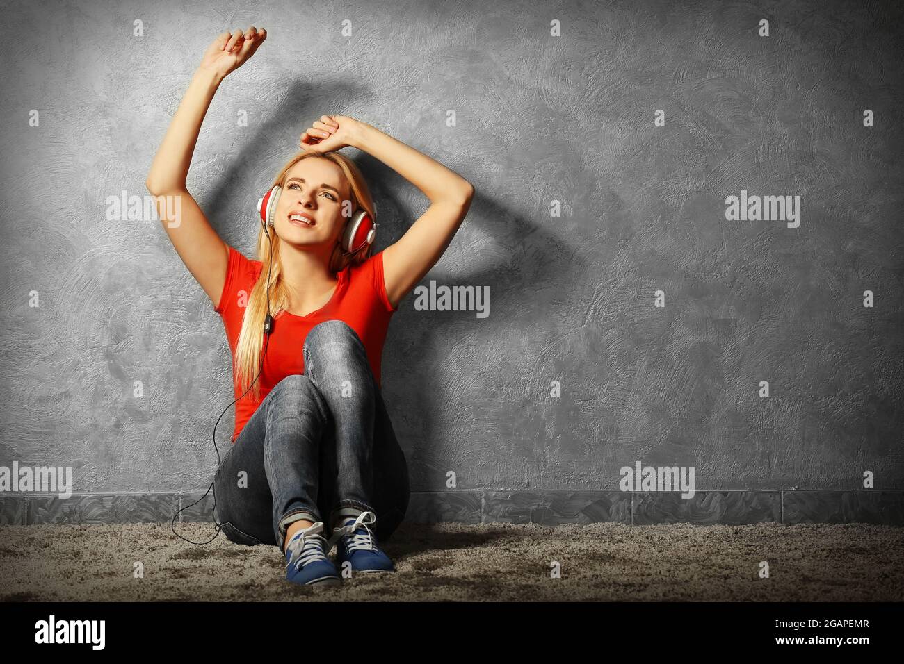 Young woman sitting on the floor and listening to music on a grey wall ...