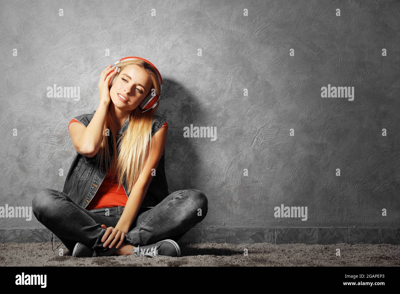 Young woman sitting on the floor and listening to music on a grey wall ...