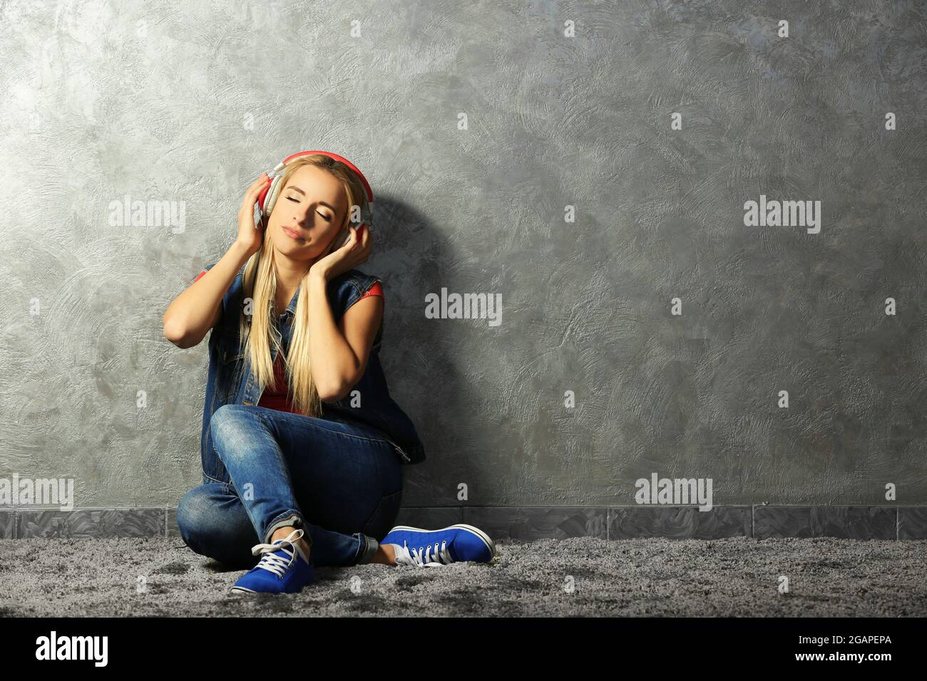 Young woman sitting on the floor and listening to music on a grey wall ...
