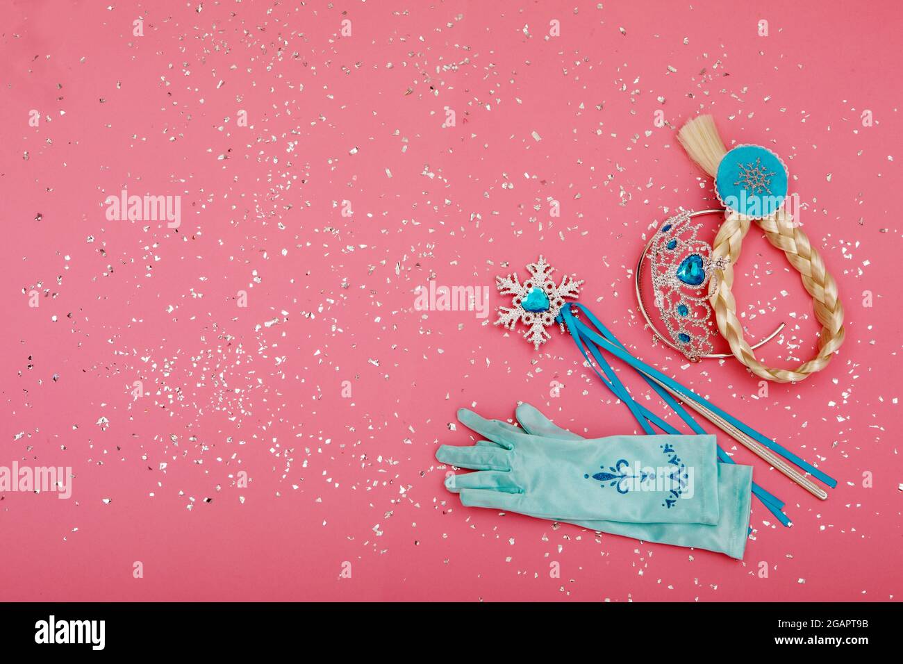 From above of magic wand and gloves placed near crown and pigtail on pink background with silver glitter Stock Photo