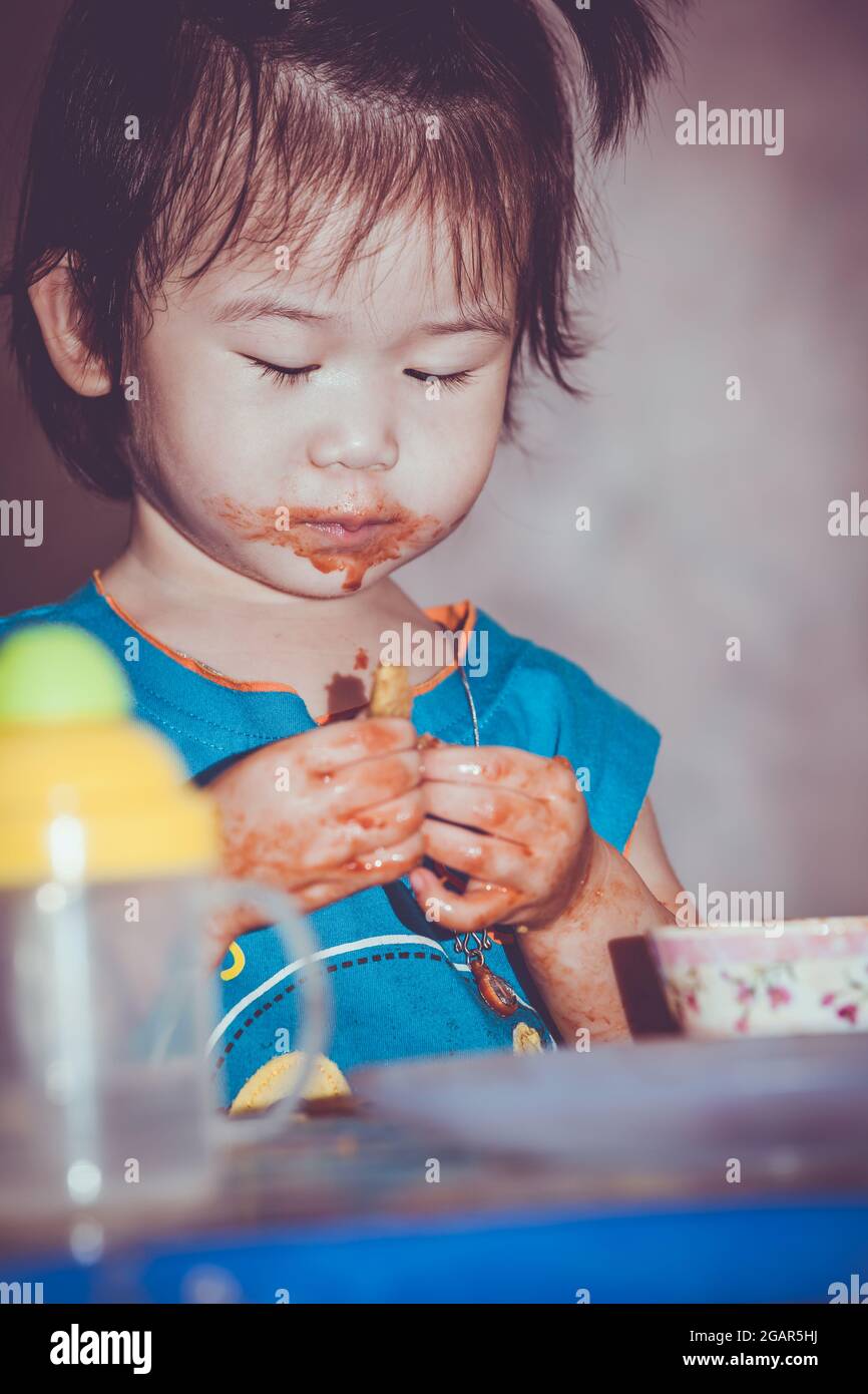 Portrait of a asian child enjoy eating. Her hand and mouth get stained with ketchup. Charming girl playing with food and learning to eat. Dirty face o Stock Photo