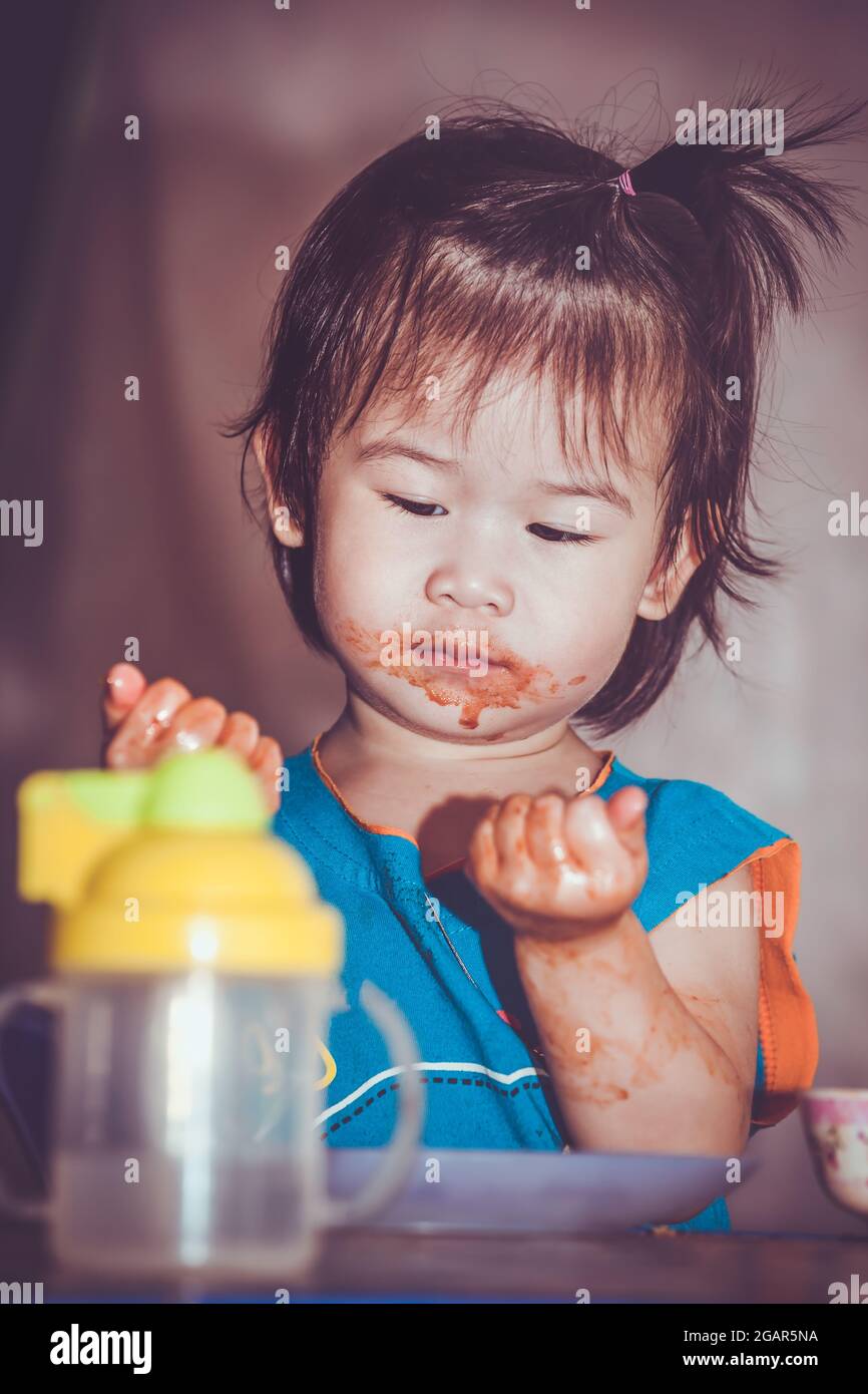 Portrait of a asian child enjoy eating. Her hand and mouth get stained with ketchup. Charming girl playing with food and learning to eat. Dirty face o Stock Photo