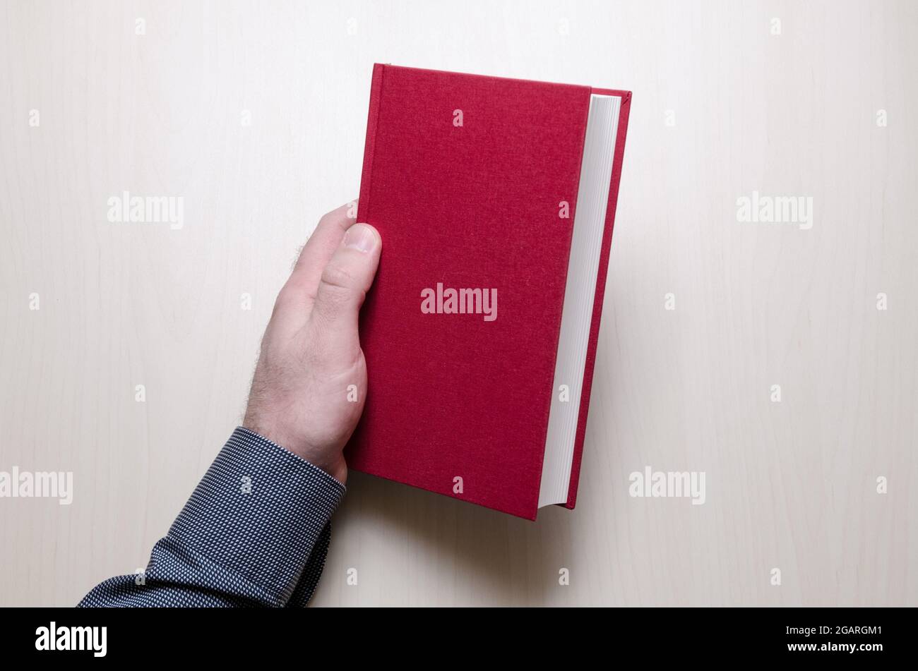 Hand holding red hardcover book or bible above wooden table or desk in one hand, indoors, literature, library concept Stock Photo