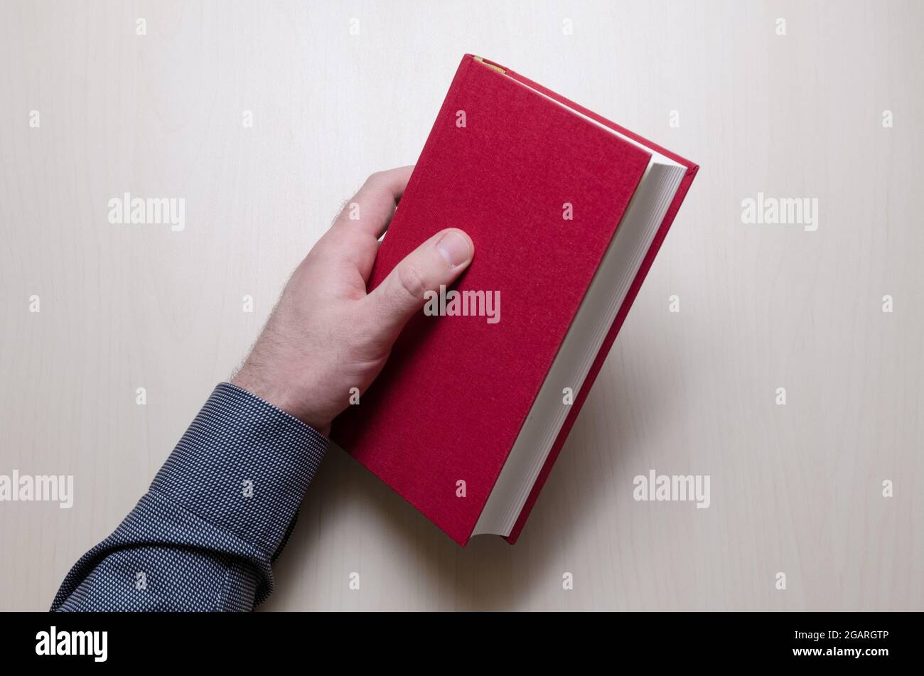 Hand holding red hardcover book or bible above wooden table or desk in one hand, indoors