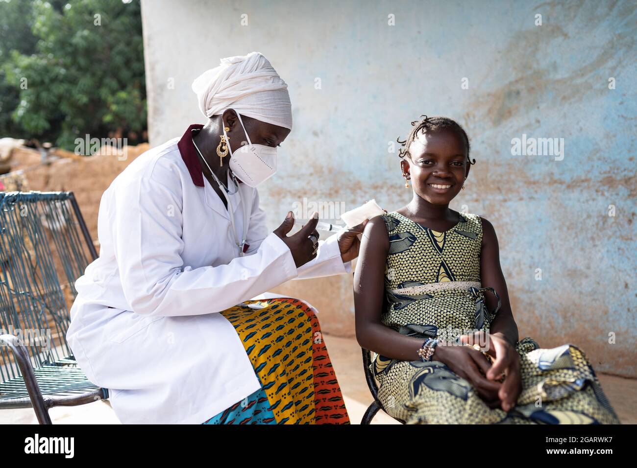 In this image, a courageous little African girl is smiling into the camera while receiving a vaccine shot by a gentle young black nurse sitting next t Stock Photo