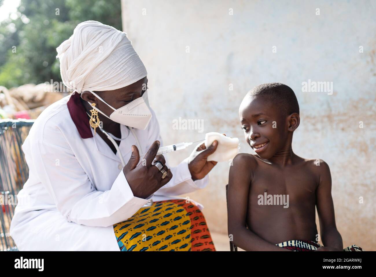 In this image a black nurse with face mask and hospital uniform is trying to explain a confident looking little African boy how she's going to inject Stock Photo