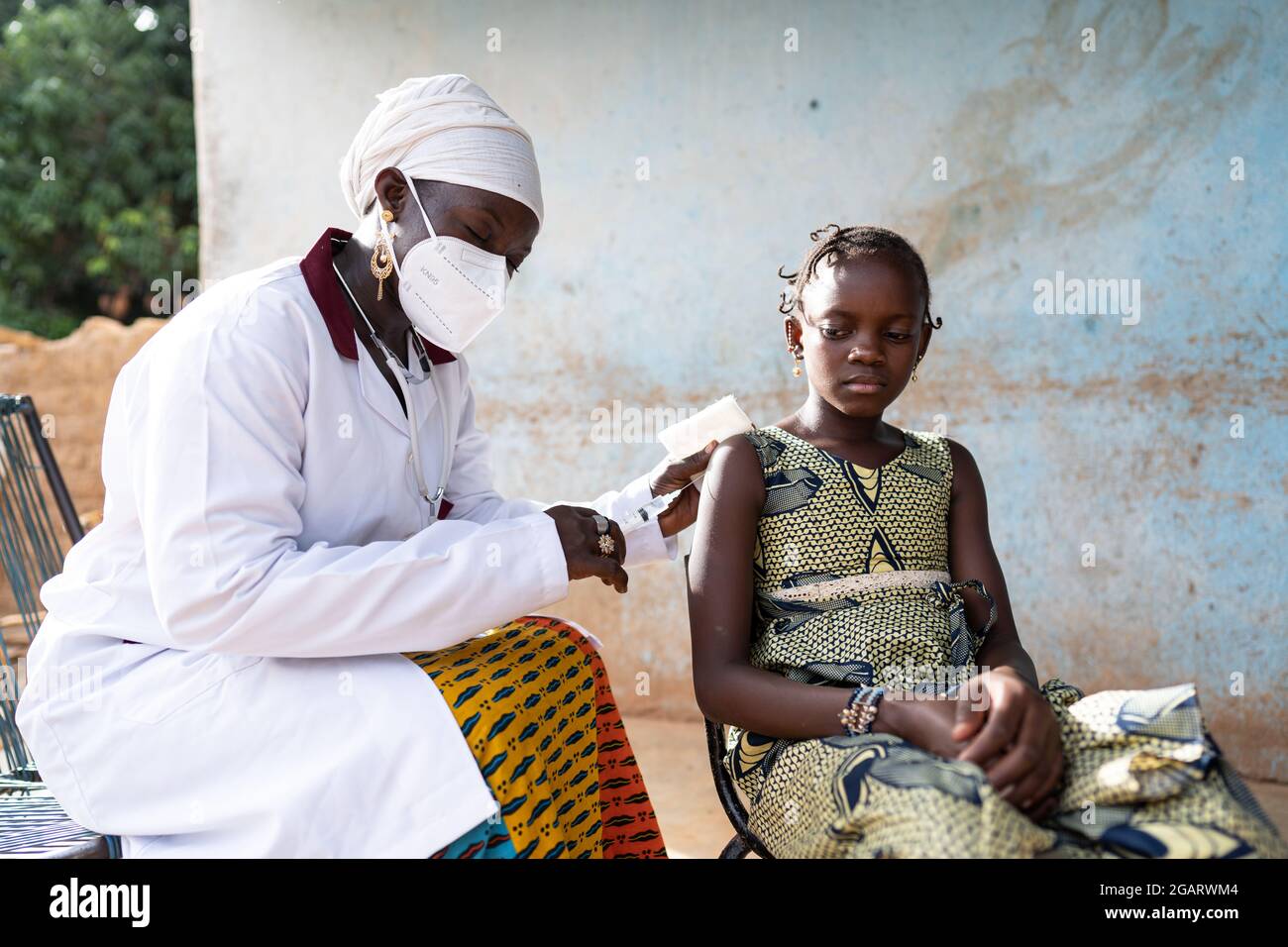 In this image, a gentle black nurse with protective face mask is delicately injecting a vaccine into the arm of a relaxed little African schoolgirl du Stock Photo
