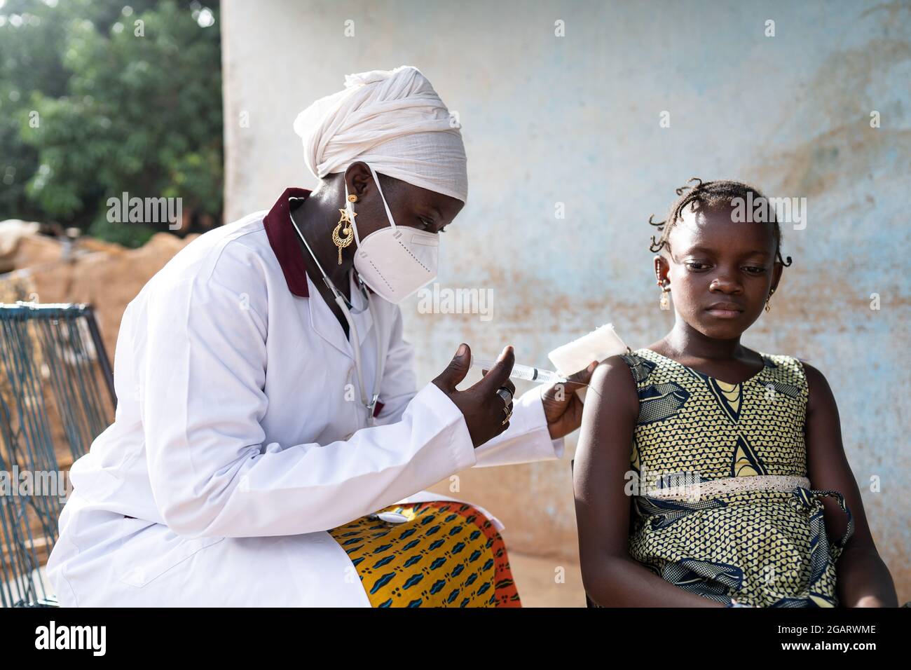 In this image, a black nurse with a big white facial mask is injecting a vaccine into the arm of an unsmiling little African girl both sitting outside Stock Photo