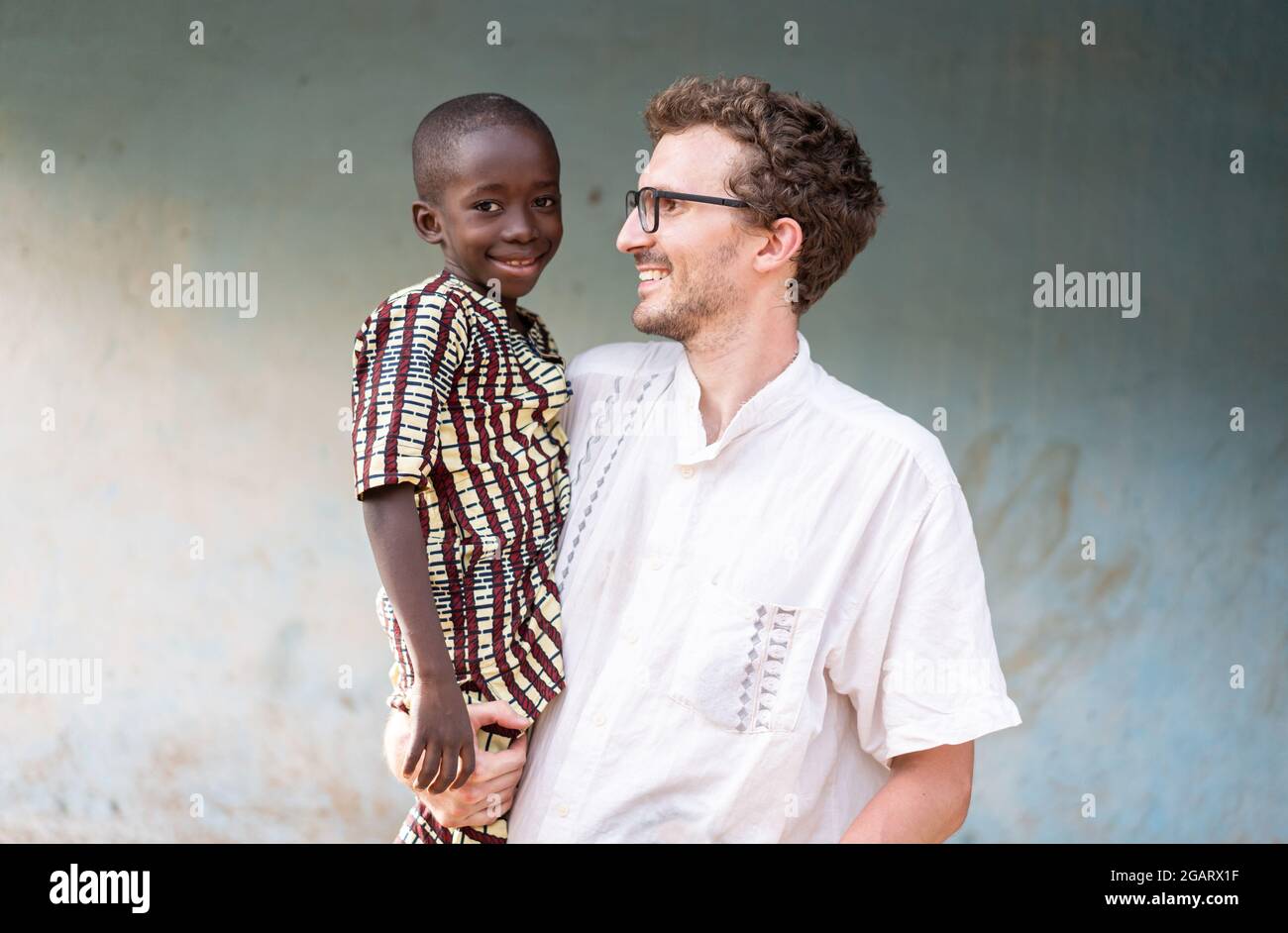 Profile view of a young caucasian man carrying a smiling little African orphan y in traditional outfit looking in camera; inernational adoption concep Stock Photo