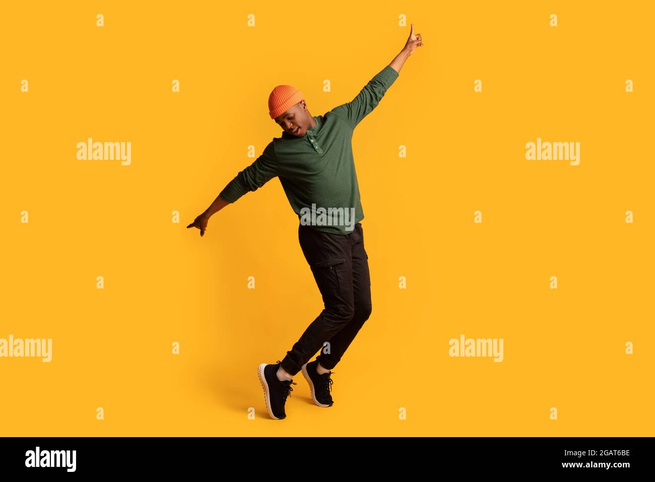 Cheerful young african american man in orange hat standing on tiptoes over yellow background, full length shot of happy millennial black hipster guy d Stock Photo