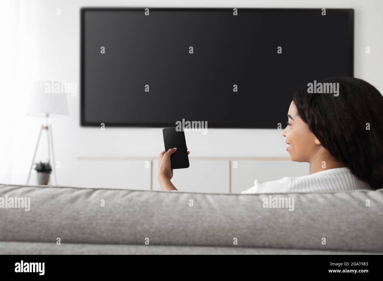 Smart home. Excited african american woman with cellphone connected to TV, plasma television set with empty black screen for mockup. Happy lady relaxi Stock Photo