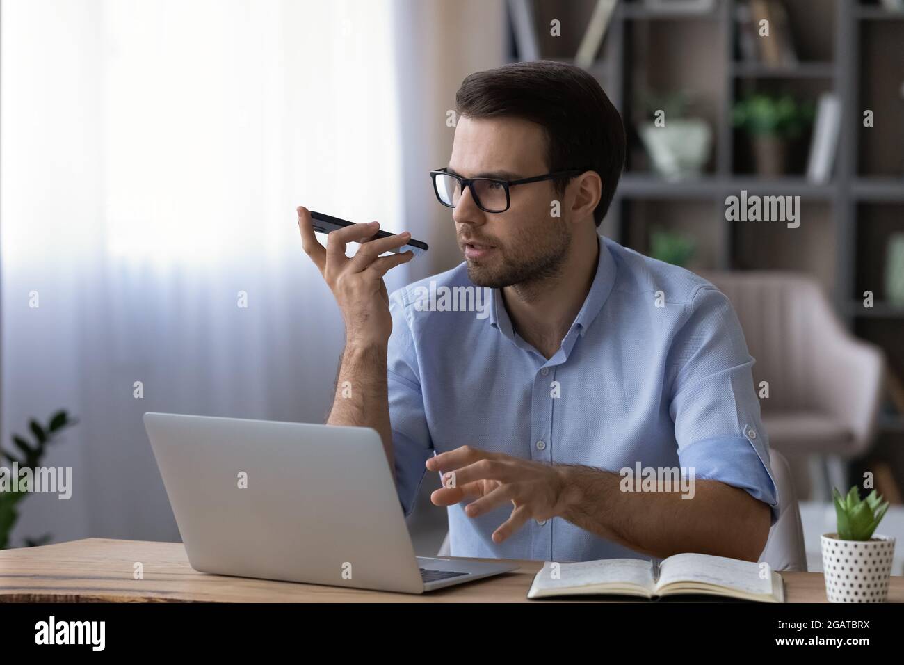 Focused male working by laptop using virtual assistant on phone Stock Photo