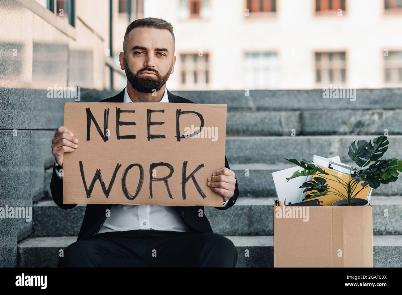 Need work. Depressed mature businessman sitting on stairs with poster and box of personal stuff, got fired due to coronavirus crisis, copy space. Unem Stock Photo