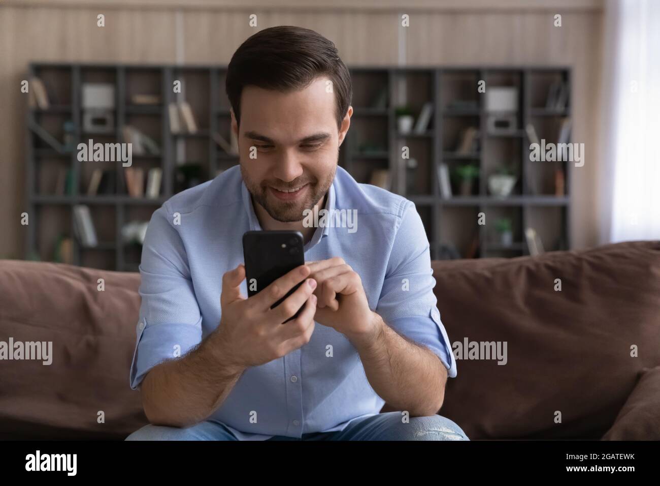 Smiling casual millennial male sit on couch chat on cellphone Stock Photo