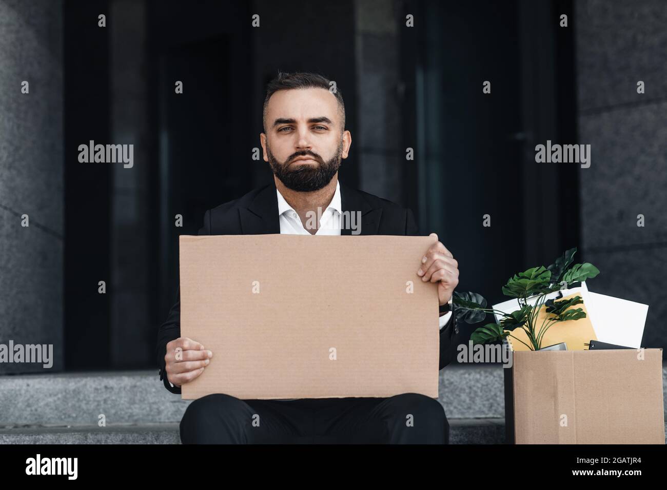 Waist up portrait of upset employee looking for job, sitting outside and holding empty poster with box of personal belongings nearby, copy space Stock Photo