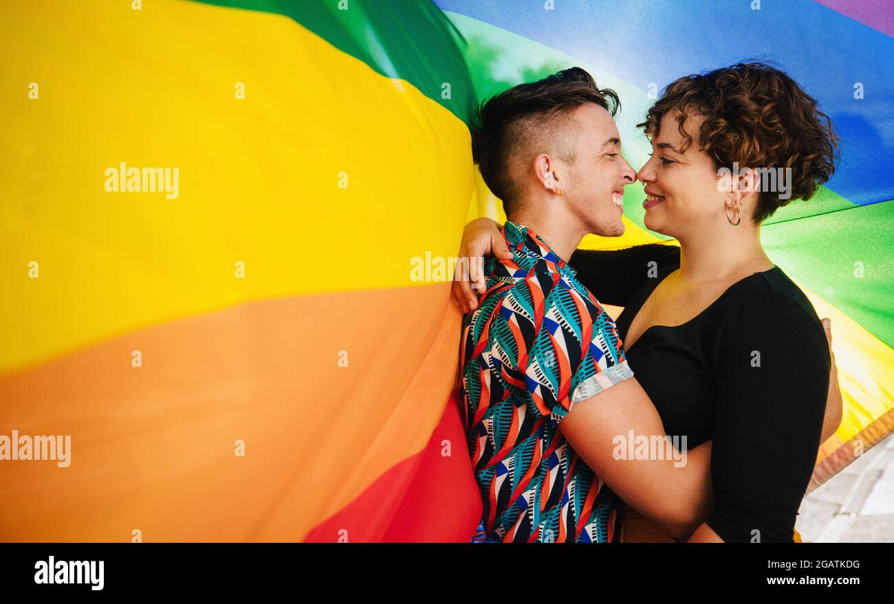 Affectionate queer couple bonding against a rainbow pride flag. Young LGBTQ couple embracing each other and touching their noses together. Two non-con Stock Photo