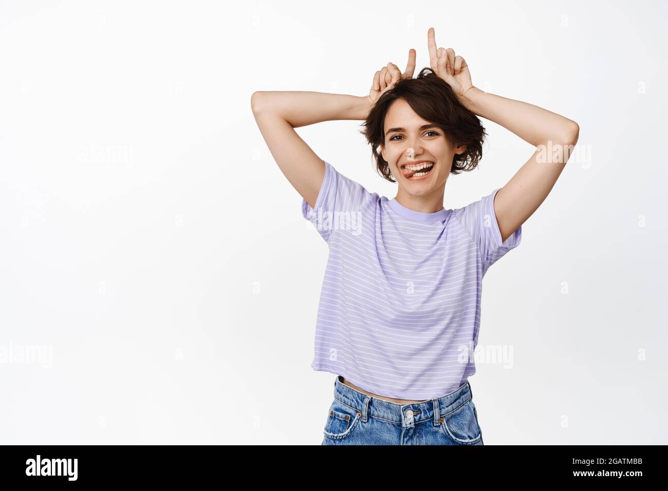 Funny and happy young woman showing bull devil horns gesture, fingers on head pointing up, smiling and laughing, standing over white background Stock Photo