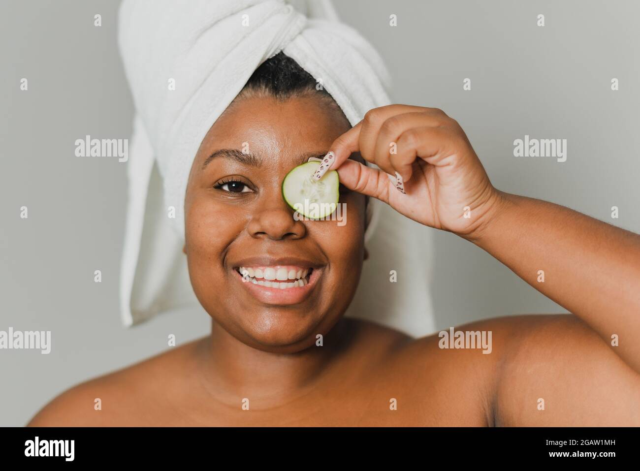 Portrait of a happy young black woman with towel taking care of her skin with a natural cucumber. Stock Photo