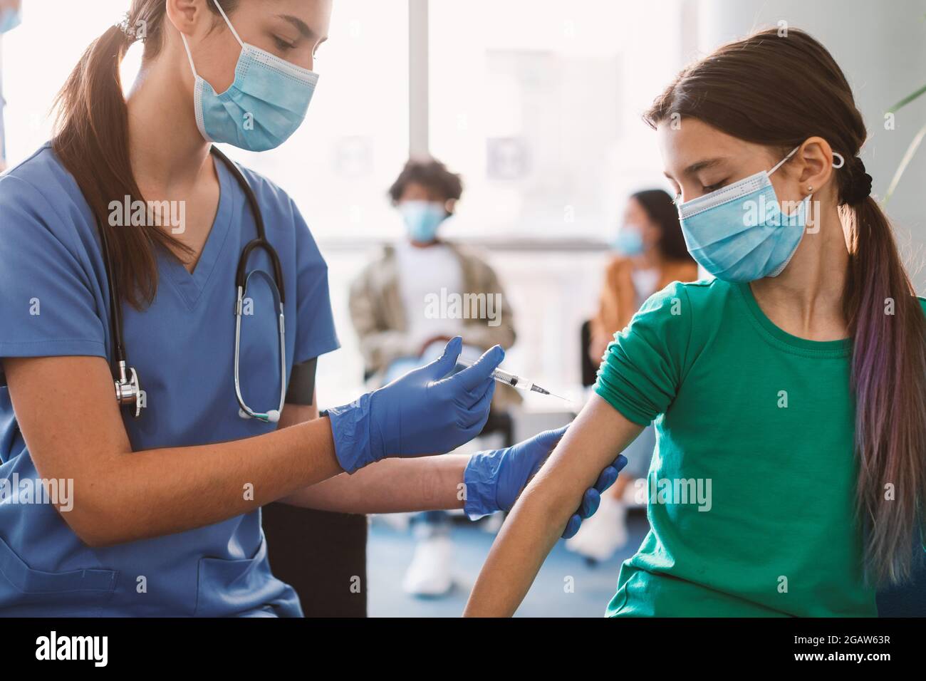 Female nurse or doctor in medical face mask giving flu injection to little patient during mass immunization campaign at health center. Schoolgirl gett Stock Photo