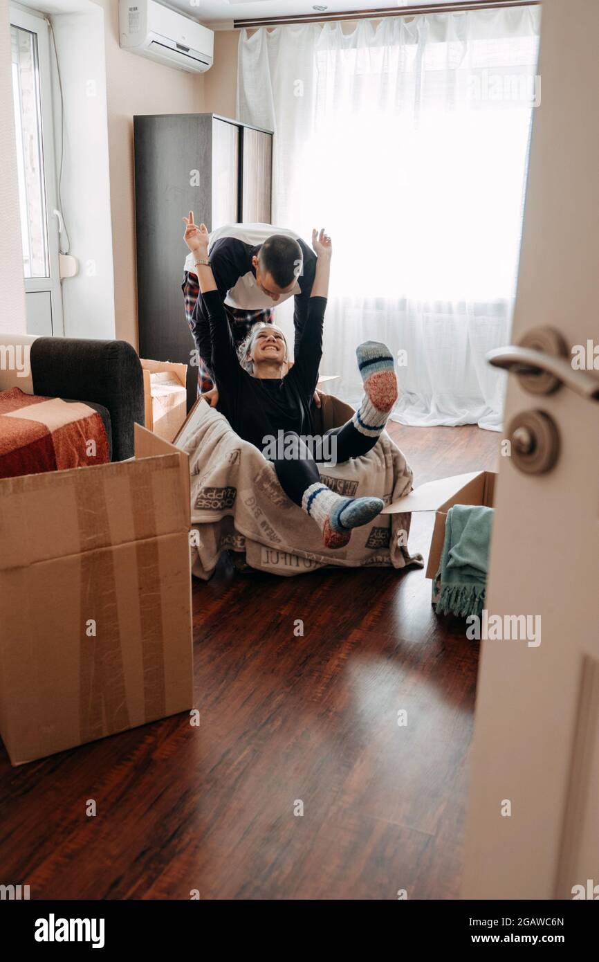 Moving day, Move Into New Home. Young happy couple, newlyweds family having fun in room with carton boxes with clothes and other things prepared for Stock Photo