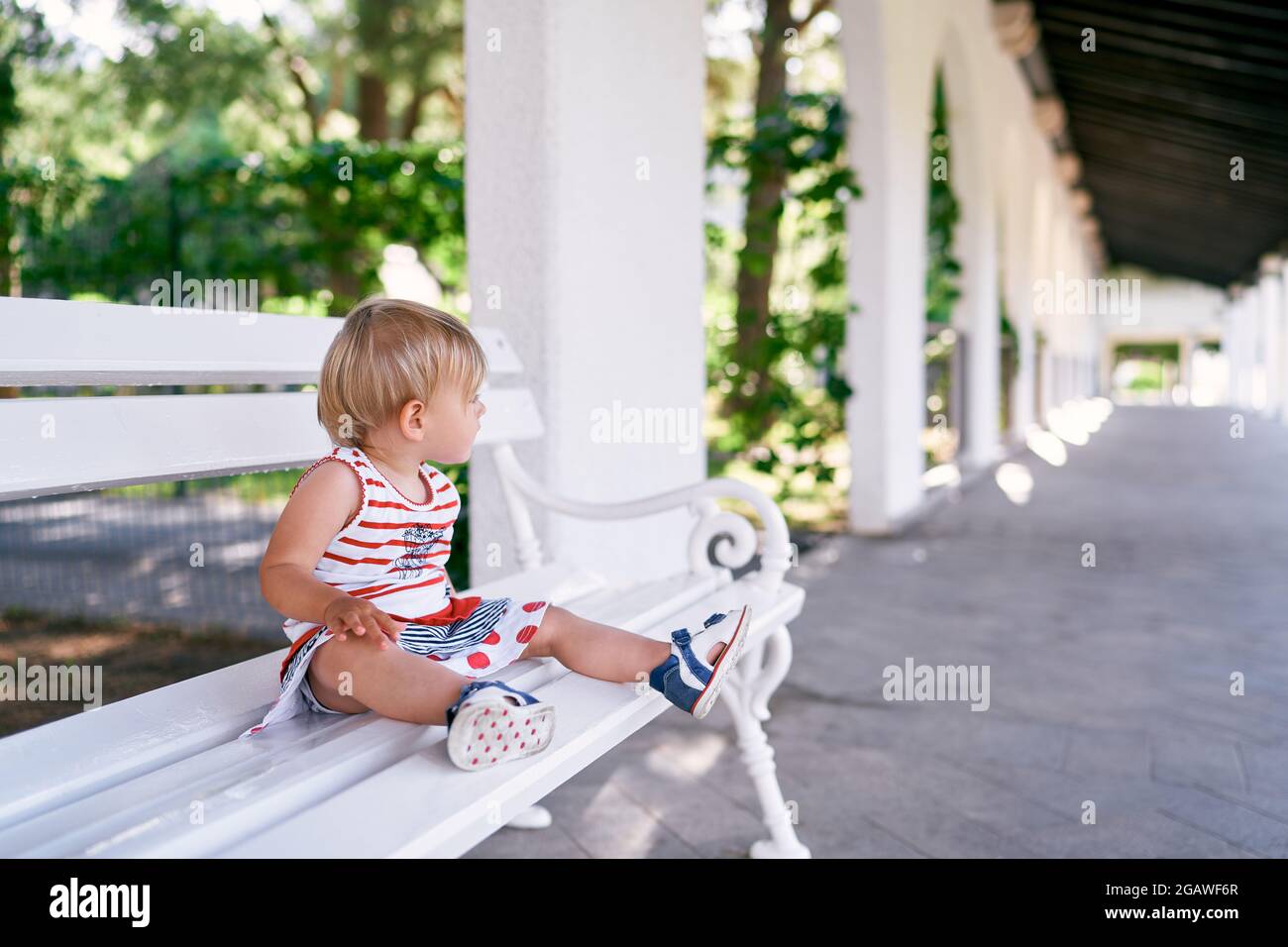 Little girl in a dress sits on a white bench and looks to the side in ...
