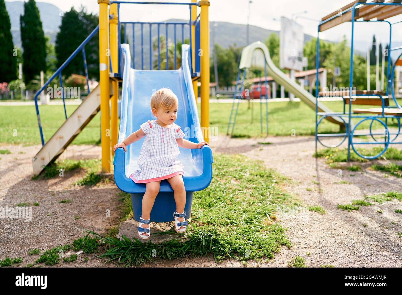 Little girl in a dress sits on a slide on the playground and looks to ...