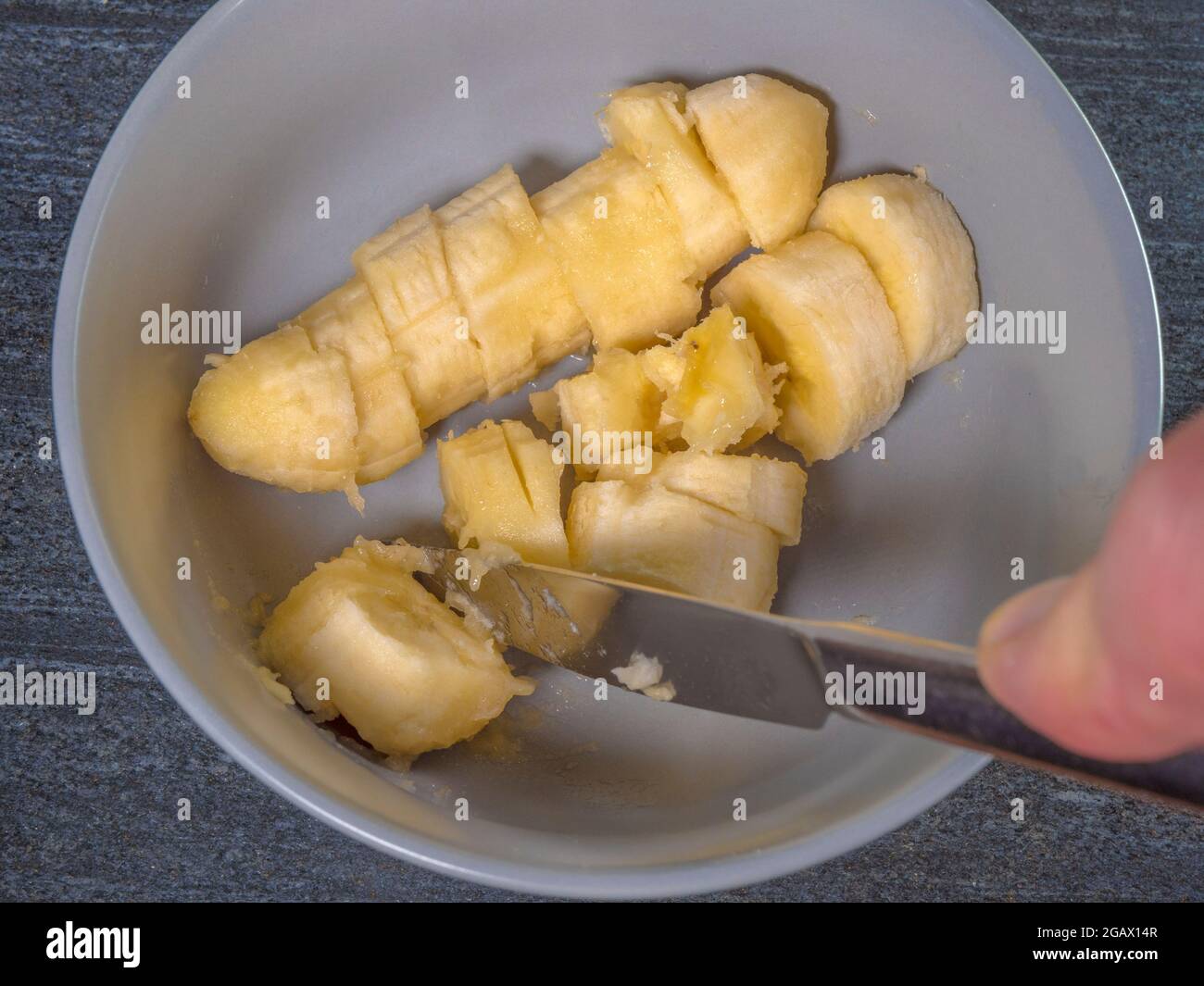 Closeup POV overhead shot of a soft ripe banana in a bowl, with the skin removed, being cut into slices with a steel knife. Stock Photo