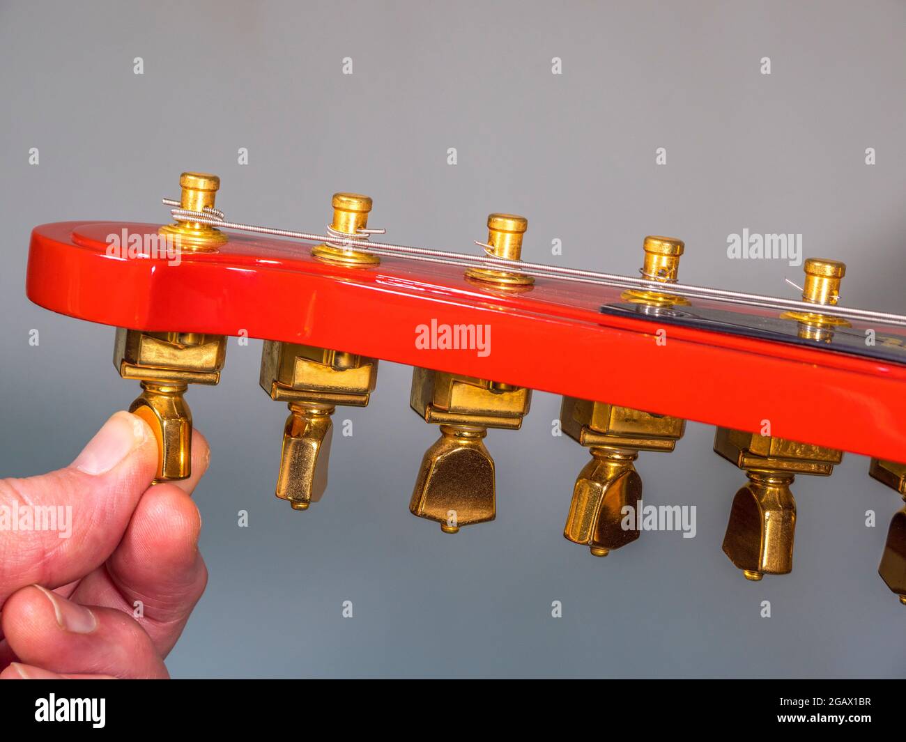 A man’s finger and thumb turning a brass tuning peg on the headstock of a bright red painted electric guitar, to loosen or tighten a wound string. Stock Photo