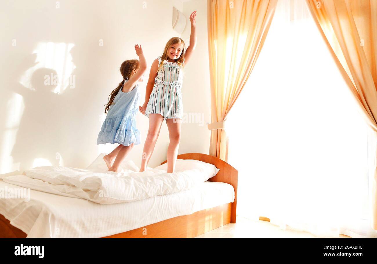 Two cute carefree little sisters girls in casual clothes playing having fun in childrens room in sunny morning. Loving joyful kids jumping on beds at Stock Photo