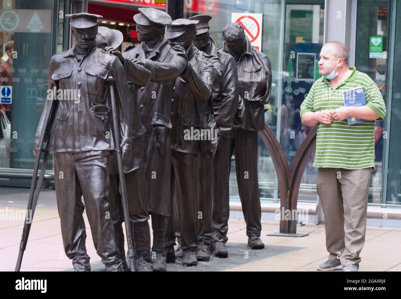 A man looks at  a statue of seven blinded soldiers outside the main entrance of Manchester Piccadilly Railway Station, central Manchester, England, United Kingdom,  Marking the centenary of the end of the First World War, it is entitled, 'Victory Over Blindness'. It was commissioned by the military charity Blind Veterans UK. The original sculpture was conceived and designed by artist and sculptor Johanna Domke-Guyot. Stock Photo