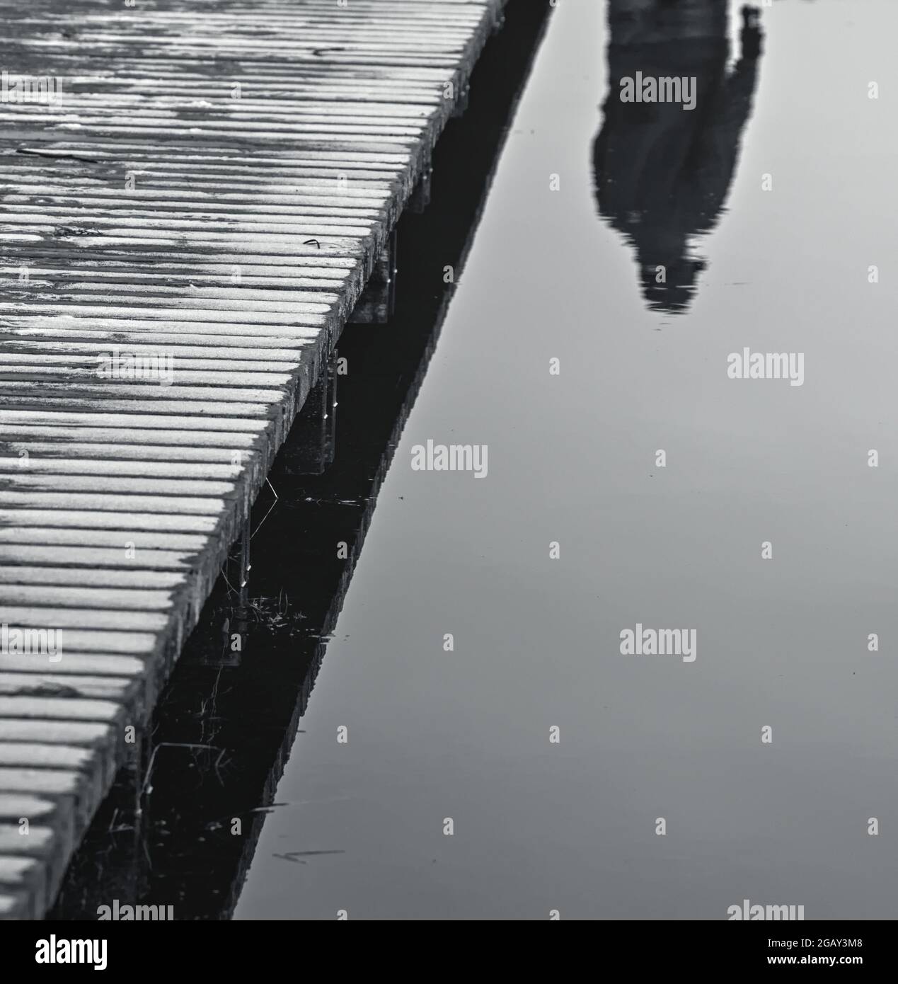 Reflection Of A Man In Monochrome Walking Across A Wooden Boardwalk Bridge Over Still Water In Winter, Christchurch UK Stock Photo