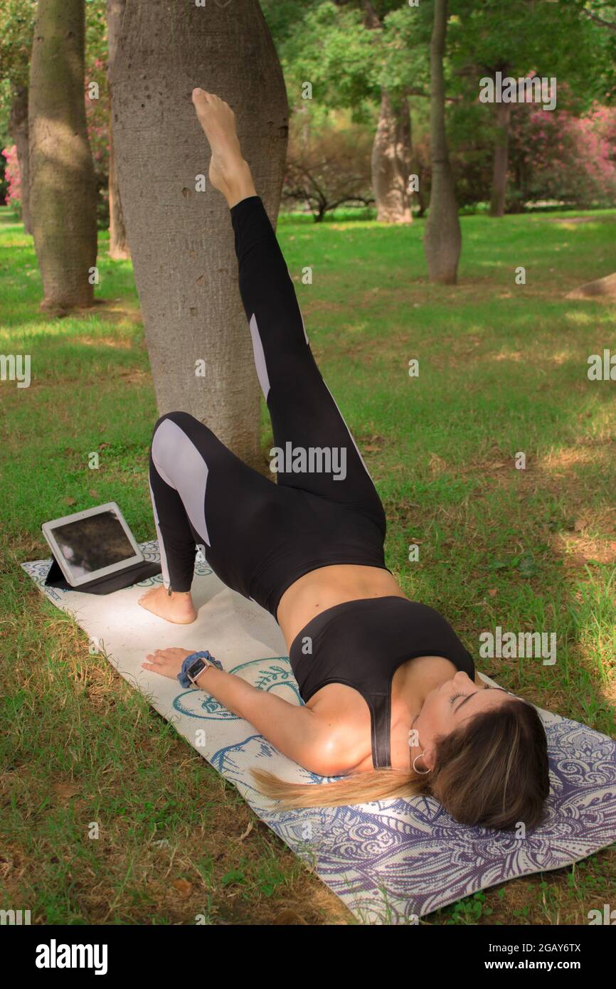 Vertical image of a young woman doing bridge yoga pose in city park in a summer sunny day. Pilates Workout Outdoor 2021. Stock Photo