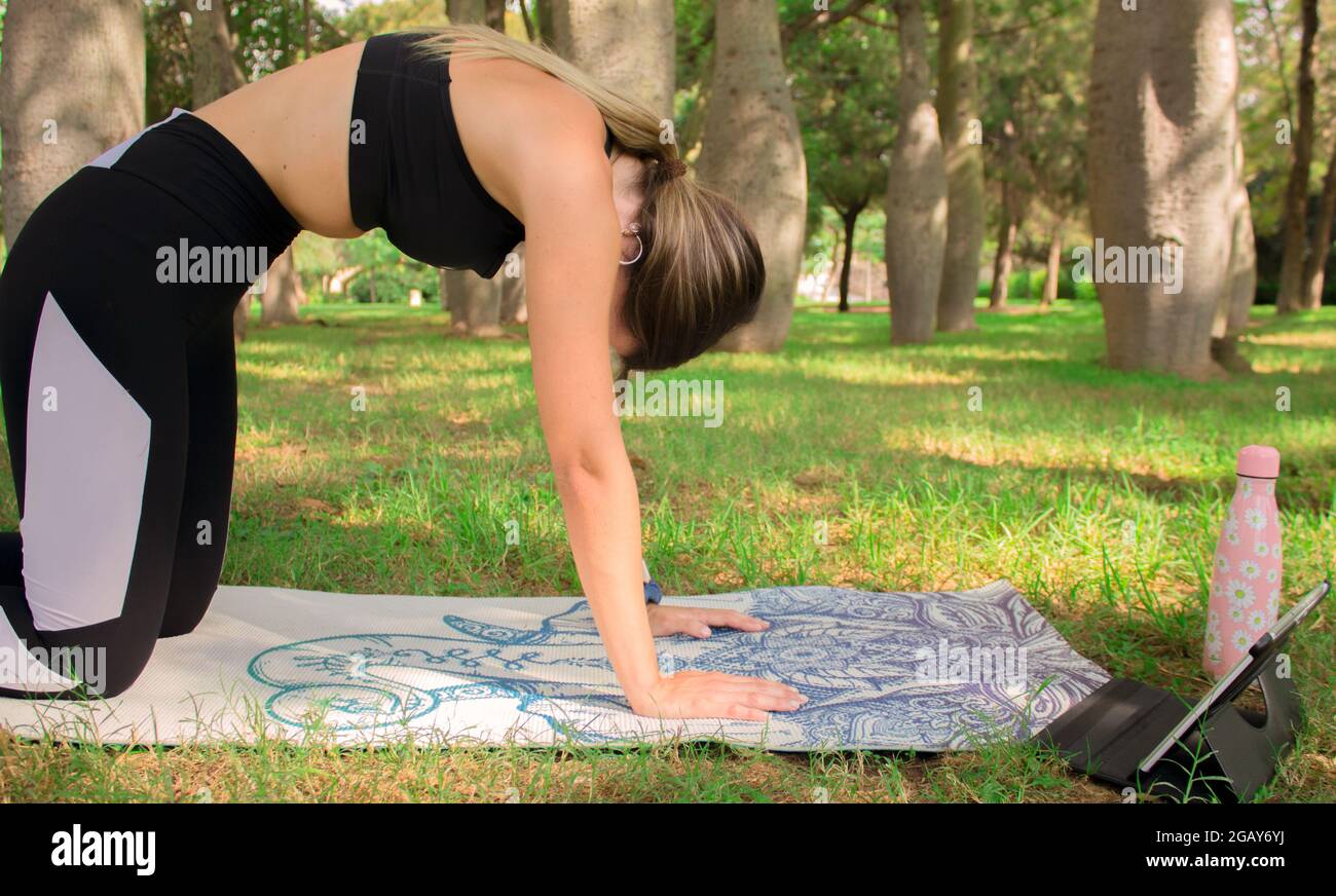 Horizontal image of a young girl doing cat yoga posture outdoor in city park in a summer sunny day.  Pilates Workout Outdoor 2021. Stock Photo
