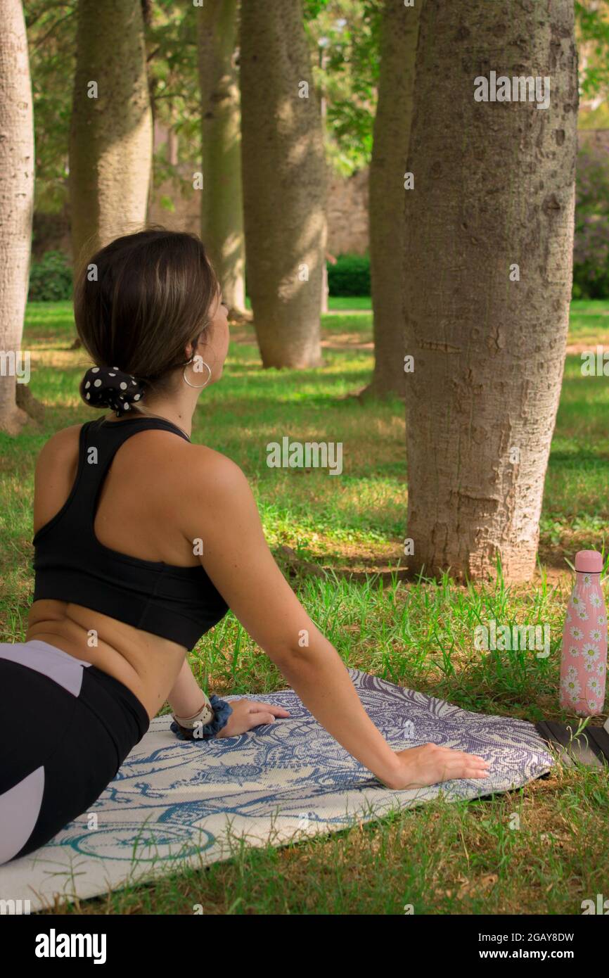 Vertical image of a young girl doing the cobra pose while she is relaxing outdoors in a summer sunny day. Pilates Outdoor 2021. Stock Photo