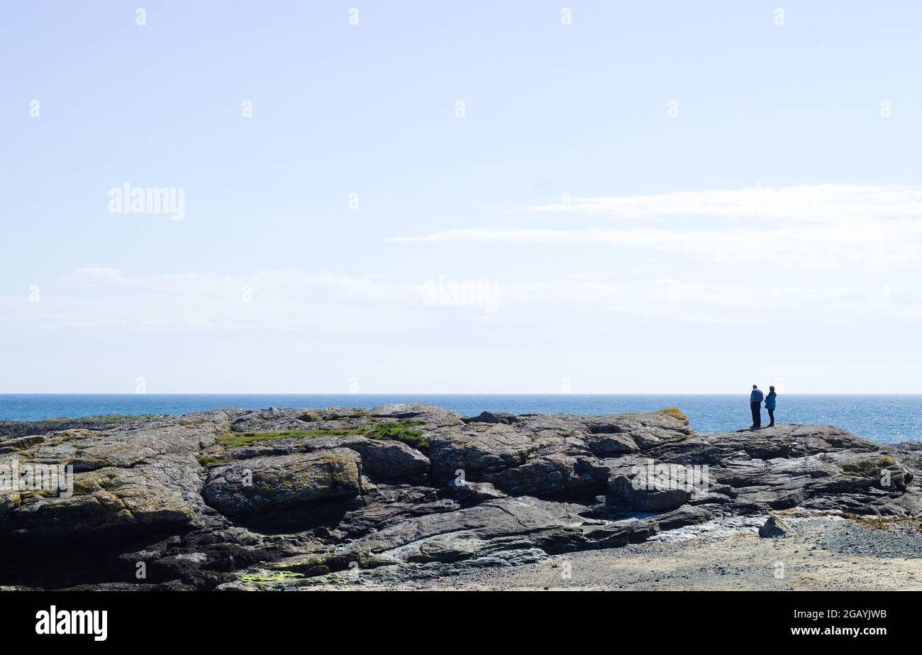 Trearddur Bay, Anglesy, Wales. Stony shoreline, holiday makers standing on low cliffs, looking out to sea. Seaside landscape. Blue sky and copy space. Stock Photo