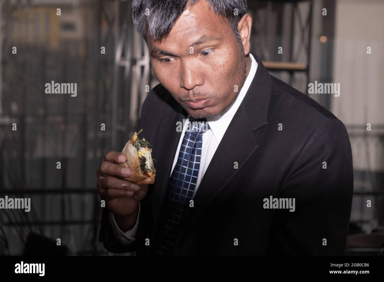 An Asian Businessman is busy with his work while eating a pizza food in the work place office but he is happy with that hard work Stock Photo