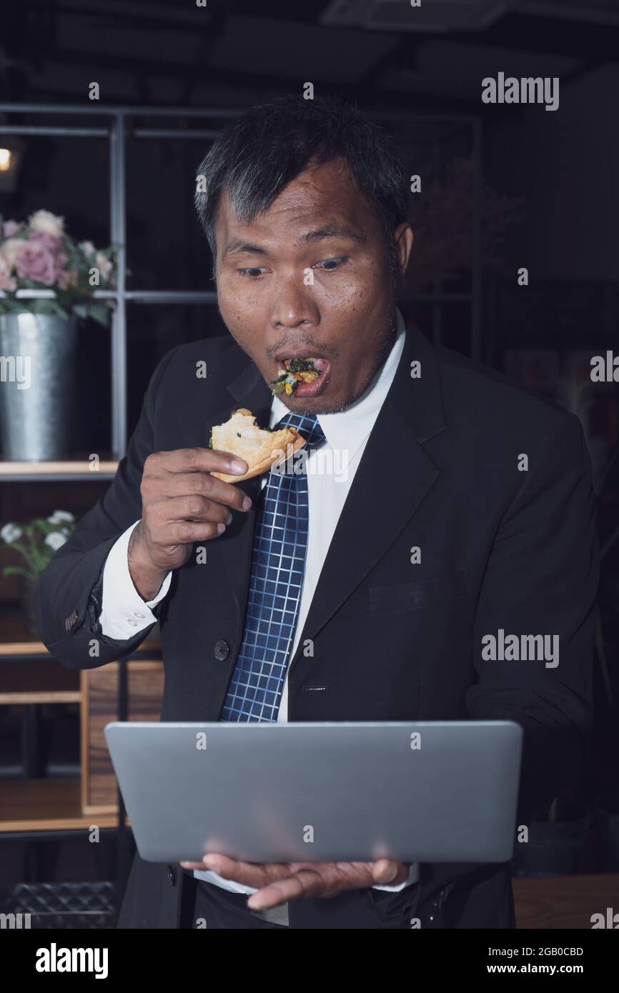 An Asian Businessman is busy with his work while eating a pizza food in the work place office but he is happy with that hard work Stock Photo